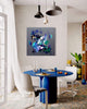 Large abstract square painting depicting a colorful, fluid and dynamic abstract composition featuring vibrant shades of blue, teal, and purple on a grey background. The painting is prominently displayed on the wall above a modern blue dining table with white chairs in a bright, airy room with pendant lighting.