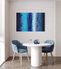 Abstract landscape painting titled "Night Illusion 80x120cm" displayed on the wall in a modern dining room setting with a white round table and blue upholstered chairs.