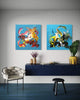 Vibrant abstract diptych paintings | Two large square abstract artwork featuring energetic, colorful brushstrokes and dynamic compositions on blue backgrounds, displayed on white brick wall in modern living room setting with dark furniture and decor.