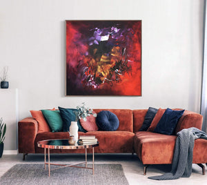 Large square abstract artwork of vibrant purple, red, and orange hues displayed on a wall above a cozy orange sofa with decorative pillows in a modern living room setting.