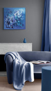 Abstract square canvas painting depicting swirling blue and white brushstrokes against a vibrant blue background, displayed in a modern living room setting with gray walls and blue accents.