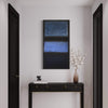 Large abstract geometric painting in blue and black tones displayed on the wall in a modern minimalist interior setting with a wooden console table and a vase of white flowers underneath.