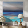 Large abstract seascape with vibrant blue and orange tones on canvas, framed in white, displayed on a wooden console table with a brass lamp and floral arrangement.
