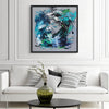 Vibrant abstract square canvas art depicting flowing blue, green, and black brushstrokes on a dark background, displayed in a modern living room setting.