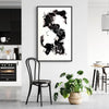 Framed abstract black and white artwork hanging on the wall in a modern home interior, featuring a dynamic, expressive brushstroke design. Potted plant in the foreground adds a natural touch to the minimalist decor.