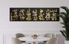 Large abstract artwork with intricate golden calligraphic strokes on a black canvas, displayed on the wall above a modern dining table with beige chairs and a potted plant in the foreground.