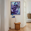 Large abstract painting featuring bold, swirling brushstrokes in shades of blue and purple, displayed on a white wall in a modern home interior with wooden floors and furniture.