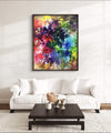 Vibrant abstract landscape painting in colorful hues of red, purple, green and yellow displayed on wall above white sofa in modern living room setting.