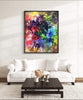 Vibrant abstract landscape painting in colorful hues of red, purple, green and yellow displayed on wall above white sofa in modern living room setting.