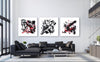 Three abstract black and white paintings depicting dynamic shapes and forms, displayed on the wall of a modern living room with a black sofa and large windows.