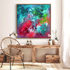 Vibrant abstract painting titled "Dancing In The Rain" depicting colorful brushstrokes in shades of blue, green, red, and pink. The abstract artwork is displayed in a wooden frame on a white wall, complementing the rustic wooden furniture and rattan chair in the room.