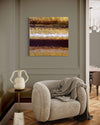 Abstract art painting on wall with textured layers of gold, brown, and white colors. Cozy gray armchair with knitted throw blanket in the foreground. Hardwood floors and wooden furnishings in the room.