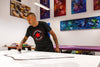 Large abstract paintings on display in an art gallery, with a man in a black Converse shirt working on a painting on an easel.