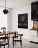 Minimalist black and white abstract painting displayed in a modern dining room setting with a wooden dining table, dark chairs, and a unique light fixture.