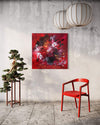 Large abstract painting with vibrant red and orange hues on a square canvas, displayed on a concrete wall alongside a bonsai tree and a modern red chair.