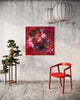 Large abstract painting with vibrant red and orange hues on a square canvas, displayed on a concrete wall alongside a bonsai tree and a modern red chair.