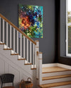 Vibrant abstract canvas painting depicting a colorful, dynamic composition with splashes of blue, green, and warm tones, displayed on the wall of a staircase in a modern home interior.