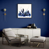 "Earthy Abstract Artwork" - Framed blue and white abstract print displayed on a blue wall above a modern white credenza, with a comfortable gray armchair and matching ottoman in the room.