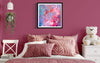 Vibrant abstract art print titled "Enchanted I" displayed on the wall of a cozy bedroom interior. The print features a colorful, expressive design in shades of pink and blue. The bedroom features soft pink bedding, a white chair, and a stuffed bear toy, creating a warm and inviting atmosphere.