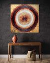 Textured abstract painting with concentric circles and warm tones displayed on a wall above a wooden table with a vase and decorative items.