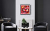 "Vibrant Abstract Art Print" - Colorful abstract painting featuring bold red, orange, and blue brushstrokes displayed in a modern black and white living room setting with comfortable armchairs and a coffee table.