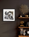 Framed abstract art print "Dancing Shadows II" displayed on dark wall with wooden shelves showcasing decorative coral and ceramic pieces