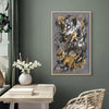 Earthy abstract artwork in a modern home interior. The large painting with brushstrokes of gray, black, and golden tones takes center stage on the sage green wall. The painting is framed and displayed above a wooden table, accented by a vase of white flowers and other decorative items. The wicker chair and plant in the room add to the natural, earthy ambiance.