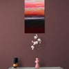 Vibrant abstract landscape artwork titled "Rothko Inspired Art | Caramel Dunes II 18x33" displayed on a burgundy wall. The painting features geometric shapes and warm tones of orange, red, and black. A delicate white floral arrangement in a pink vase sits on a table below the artwork, complementing the abstract style.