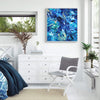 Large abstract seascape painting with vibrant blue hues and textured brushstrokes displayed on the wall of a modern, minimalist bedroom. The painting complements the natural greenery visible through the window, creating a serene and calming atmosphere.