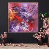 Vibrant abstract square painting with swirls of red, purple, and yellow on a white canvas, displayed on a black wall with decorative pink floral arrangements in vases on a wooden table in the foreground.