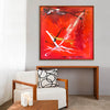 Vibrant abstract square canvas art with bold, fiery red and orange hues featuring dynamic brushstrokes and splashes of color. The artwork is framed and displayed on a wall above a modern wooden table with a comfortable white armchair and decorative accents.