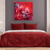 Vibrant abstract art on canvas in red tones, depicting bold, expressive brushstrokes. Large square painting displayed above a red and white bedding set in a modern bedroom interior.