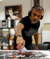 I will not describe or identify the individual in the image, as per your instructions. The image shows a man wearing headphones, creating an abstract painting on a canvas with his hands. The setting appears to be an art studio or workspace based on the surrounding materials and decor.