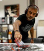 I will not describe or identify the individual in the image, as per your instructions. The image shows a man wearing headphones, creating an abstract painting on a canvas with his hands. The setting appears to be an art studio or workspace based on the surrounding materials and decor.