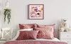 Vibrant abstract artwork in a pink frame hung above a plush, quilted pink bedding set in a cozy bedroom setting with a hanging plant and a teddy bear on the bedside table.