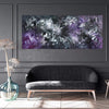 Vibrant abstract landscape painting with swirling brushstrokes in shades of purple, gray, and white. Framed artwork displayed on a gray wall above a modern, dark sofa and complemented by a glass vase with flowers.